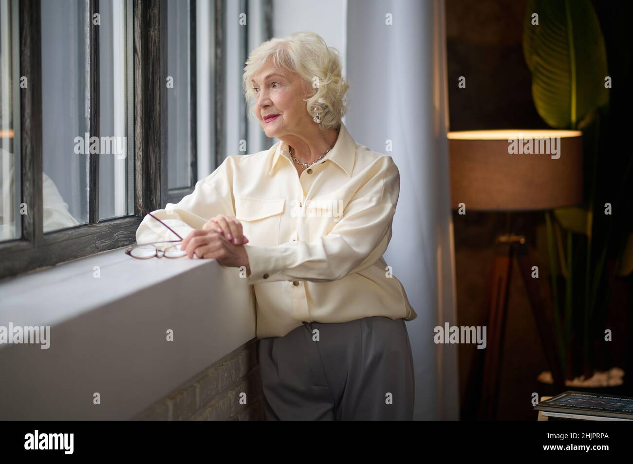 Elderly woman standing near window indoors Stock Photo