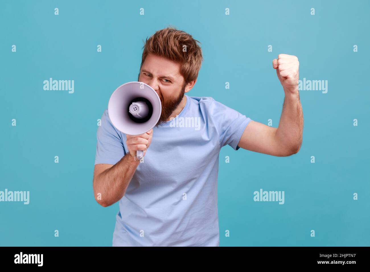 Portrait of aggressive bearded man holding megaphone near mouth, loudly speaking, screaming, making announcement with raised arm, protesting. Indoor studio shot isolated on blue background. Stock Photo