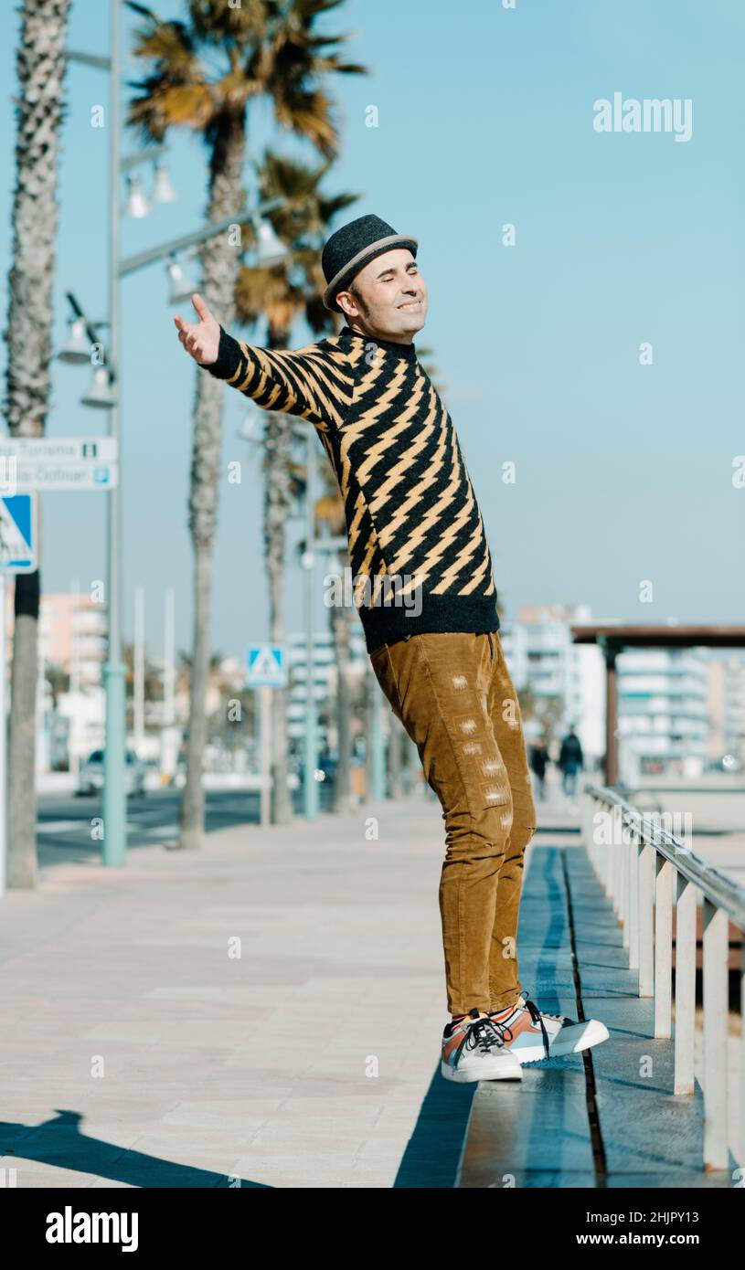 a smiley middle-aged man, wearing hat and modern casual clothes, is about to fall backwards from the bench where is standing in the promenade next to Stock Photo