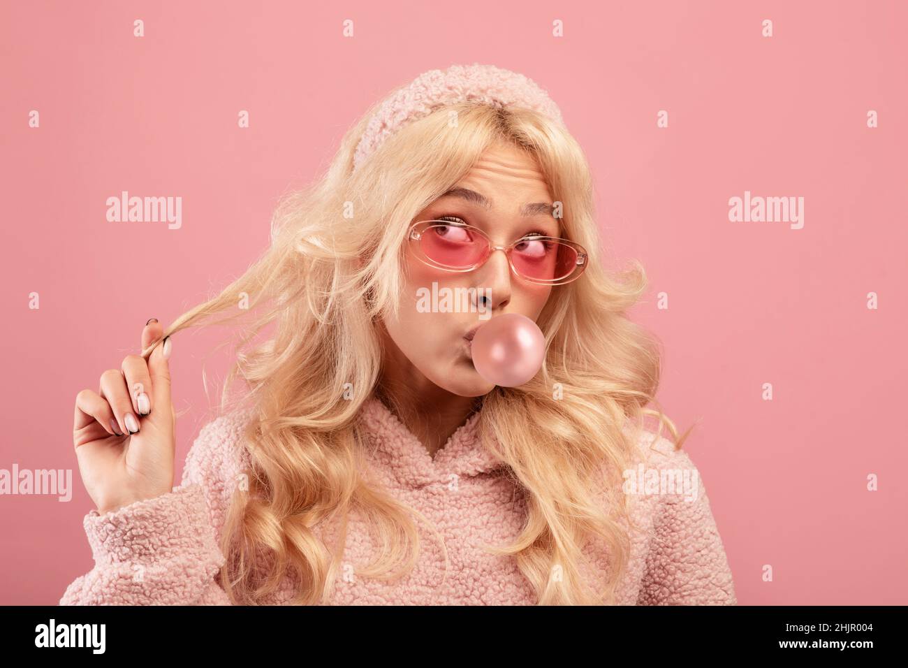 Pretty blonde lady blowing bubble from chewing gum and twisting lock of hair  on her finger, pink background Stock Photo - Alamy
