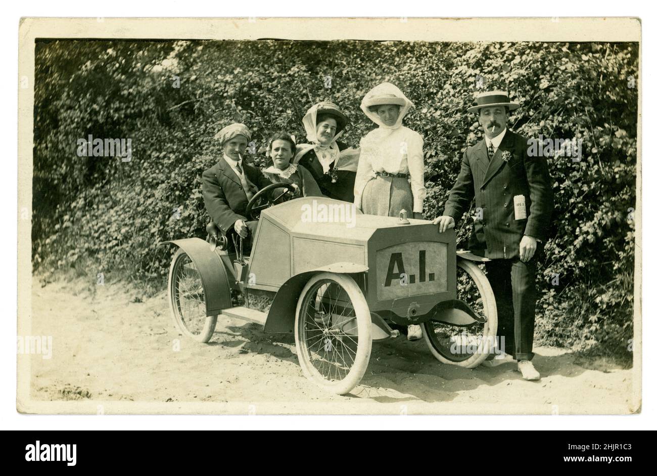 Original amusing Edwardian era postcard of happy tourists, men and women wearing straw boaters and hats, dressed in smart Sunday Best clothes, posting for a photo in a model car, photographic (some characters) prop, on a summer seaside holiday, photographed by  H. Batty, The Jetty Studio, Clacton-on-Sea, Essex, U.K. circa 1910, 1912. Stock Photo