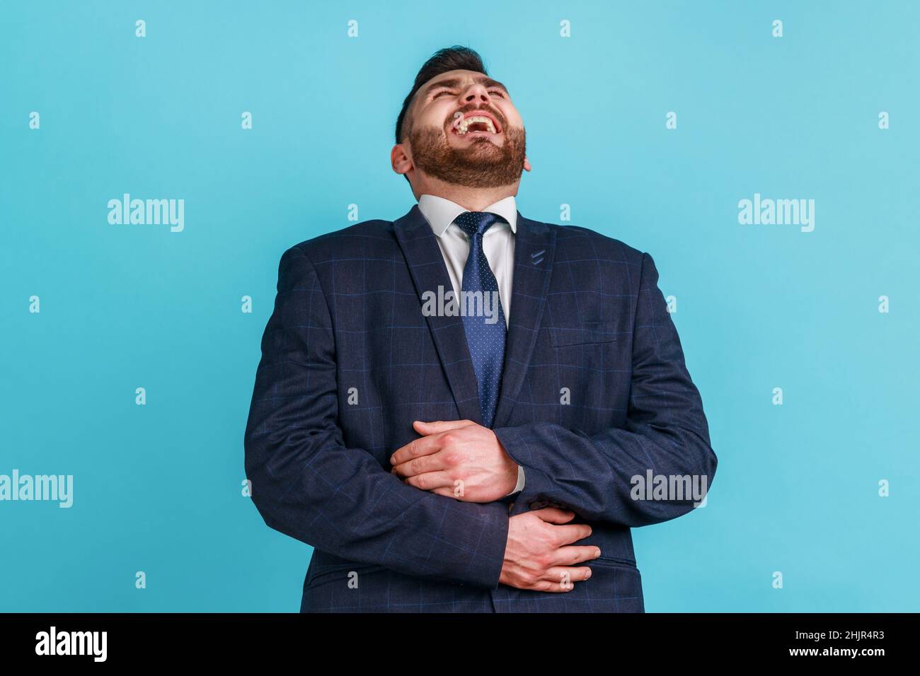 Positive bearded man wearing official style suit holding his belly and laughing out loud, chuckling and hysterically laughing with anecdote, having fun. Indoor studio shot isolated on blue background. Stock Photo