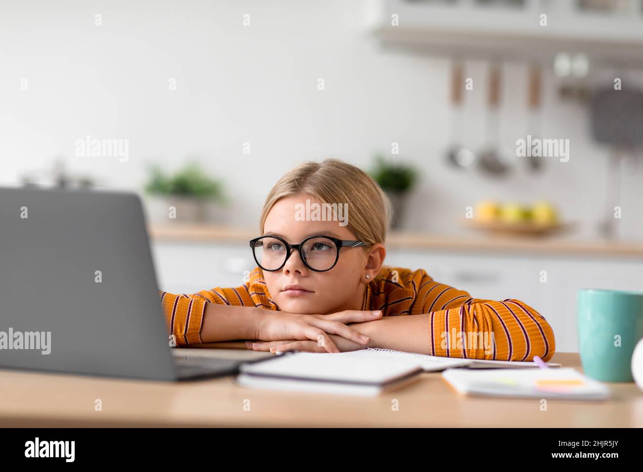 Tired serious pretty european teenage girl pupil in glasses looks at laptop, sits at table Stock Photo