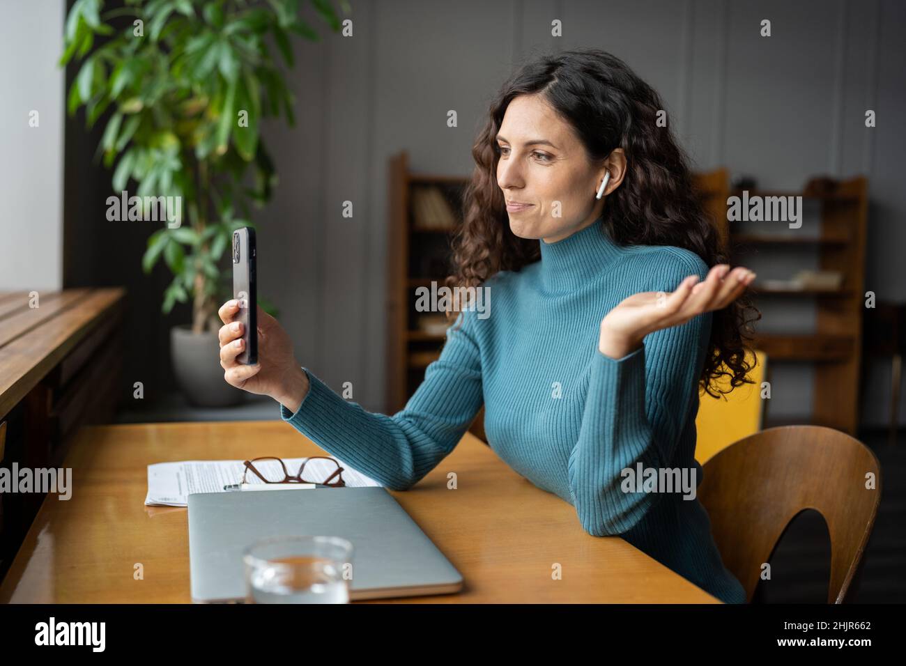 Smiling young businesswoman in earbuds sitting at office desk talking by video call on smartphone Stock Photo