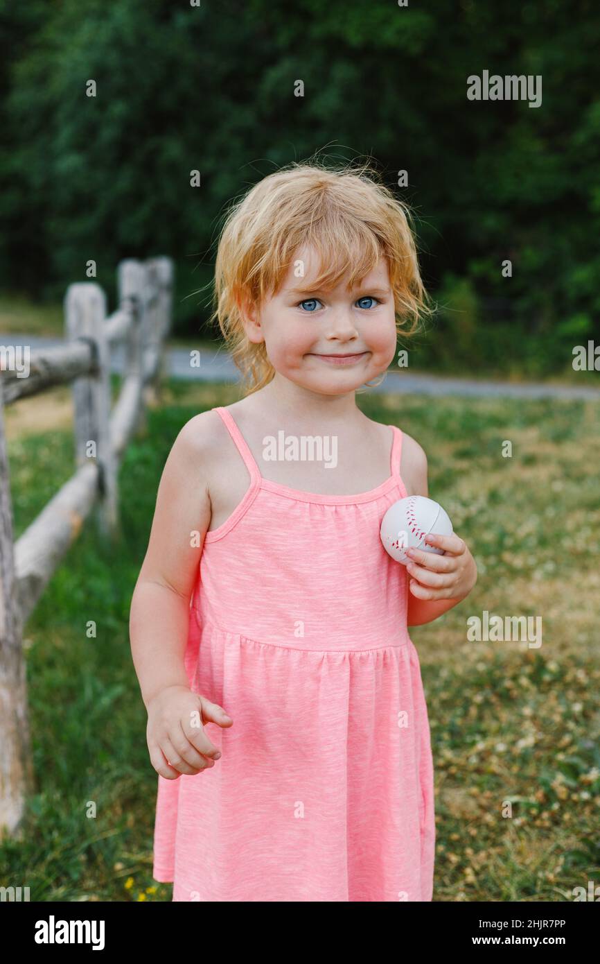 Happy little child playing with ball in park. Smiling beautiful small girl outside in summer wearing dress, having fun Stock Photo