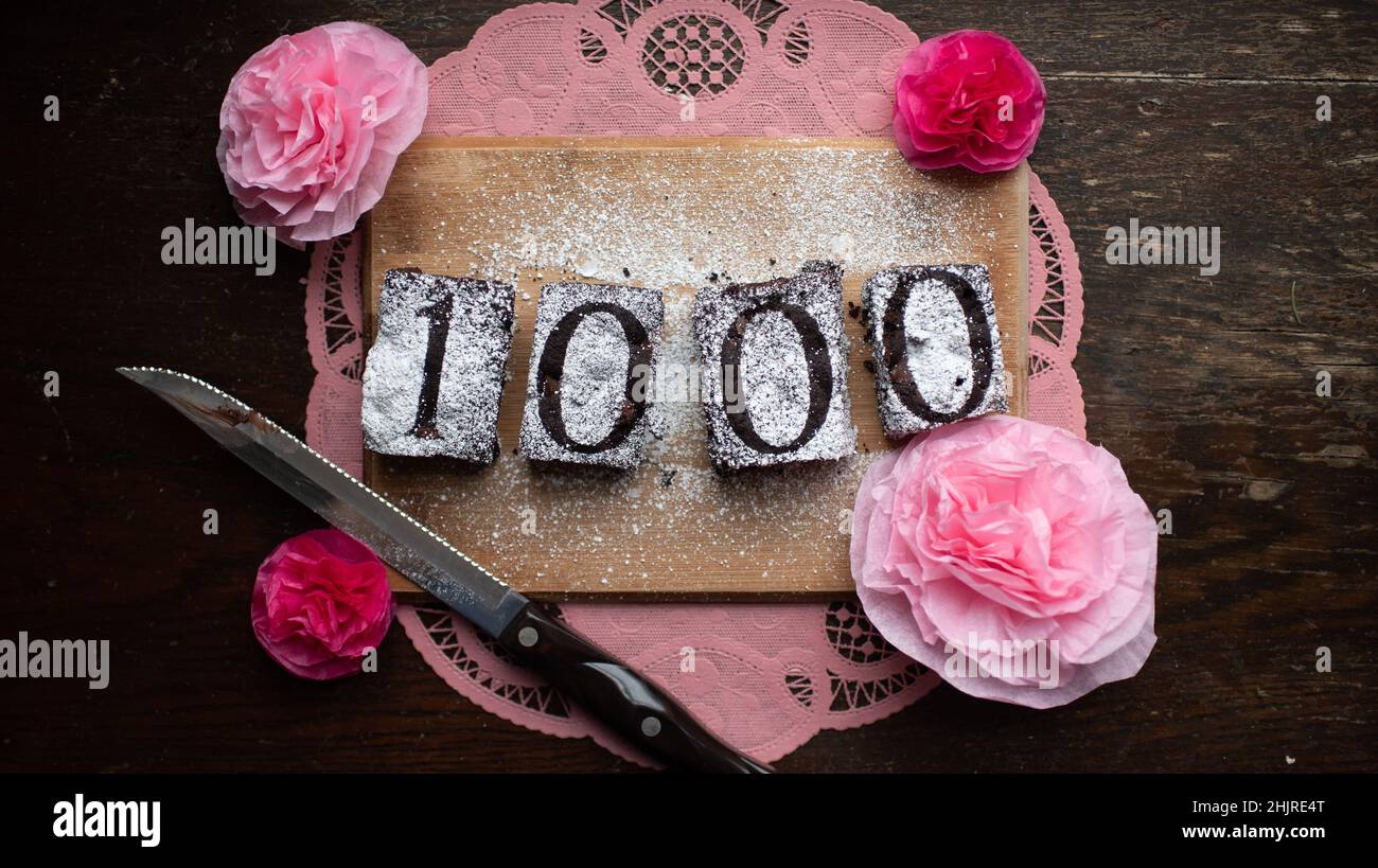 The number 1000 spelled out stenciled on chocolate brownies by powdered sugar, knife surrounded by paper pink flowers, crafted, celebration Stock Photo
