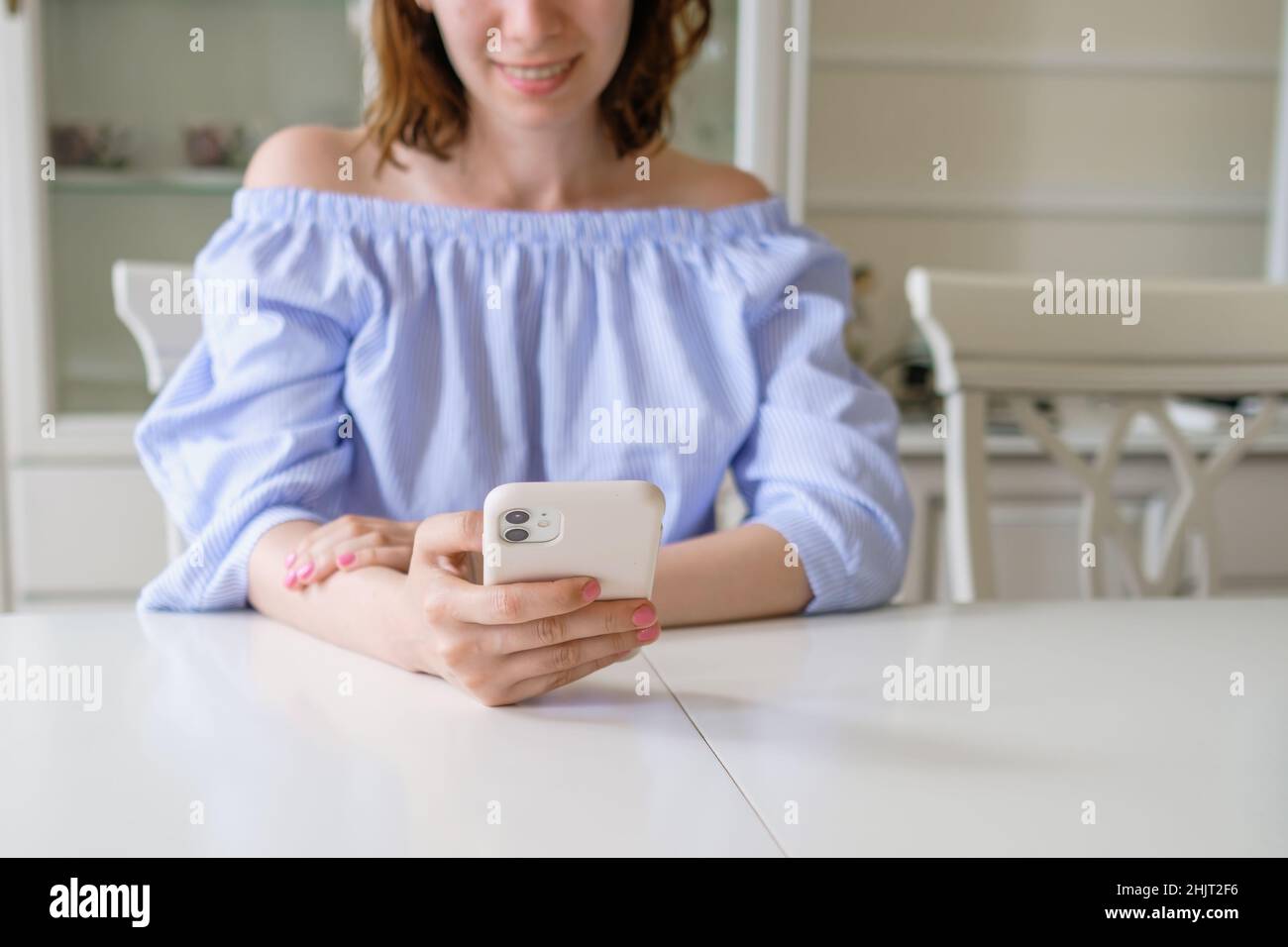 Young happy brunette woman in stylish blouse checks mail in mobile phone sitting at white table in early morning at home close view Stock Photo