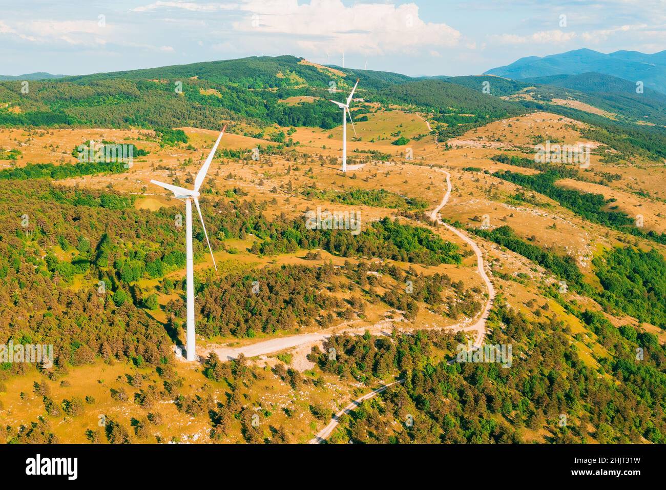 Windmills with large blades operate placed in forestry mountains producing clean energy under cloudy sky on summer day view from drone Stock Photo