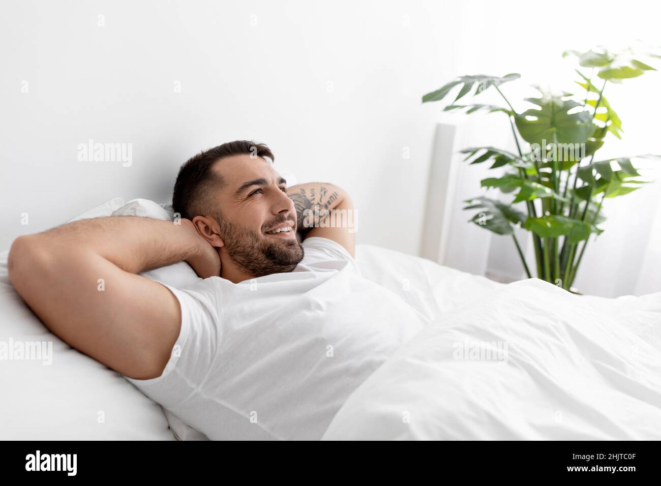 Happy caucasian young male in white t-shirt rests and relaxes, looks at empty space on bed in bedroom interior Stock Photo