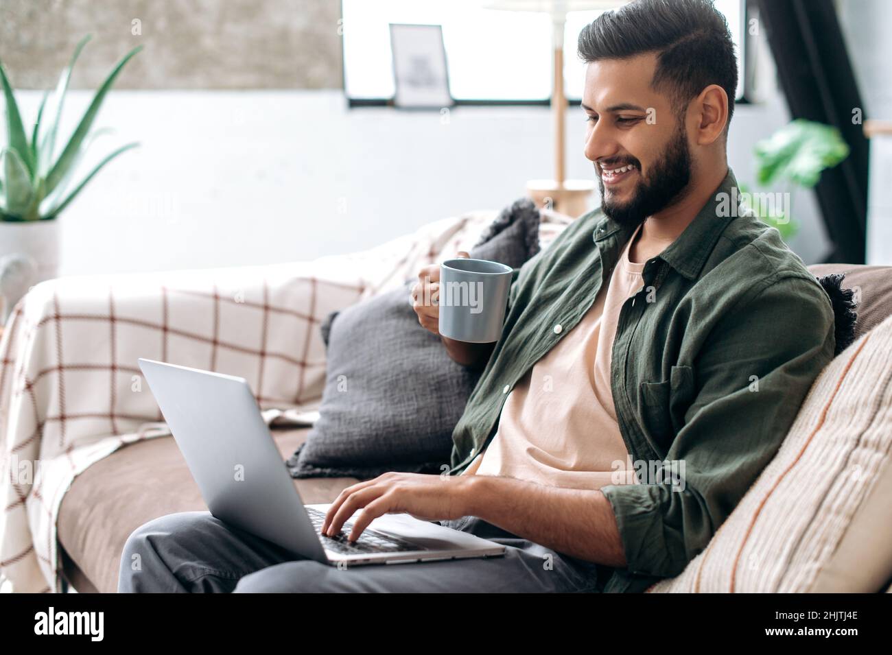 Positive handsome young indian man in casual clothes, sitting on the couch at home in the living room, using a laptop, browsing the Internet, messaging on social networks, looking at screen, smiles Stock Photo