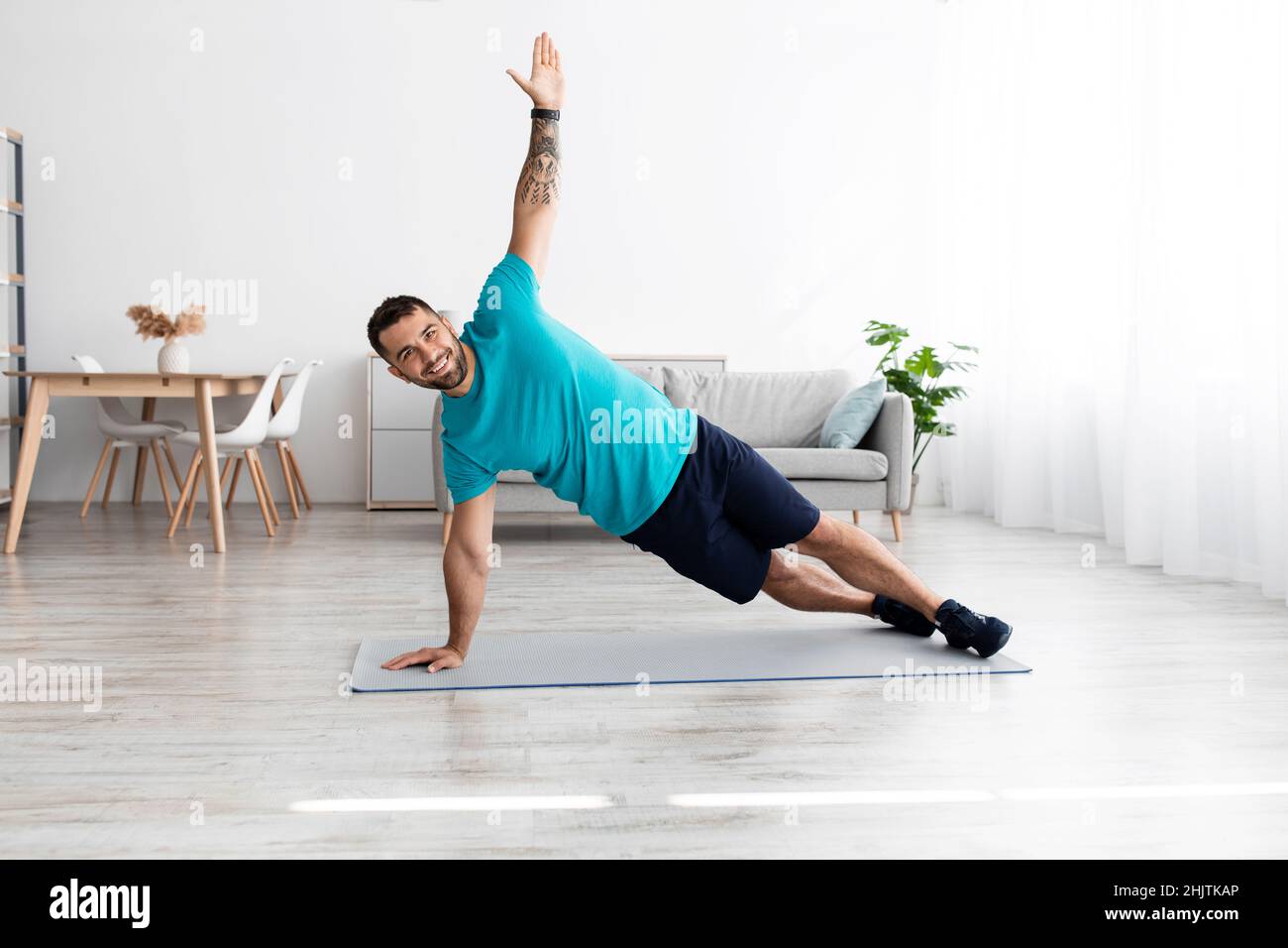 Smiling cheerful millennial european male athlete doing side plank at mat in minimalist living room Stock Photo