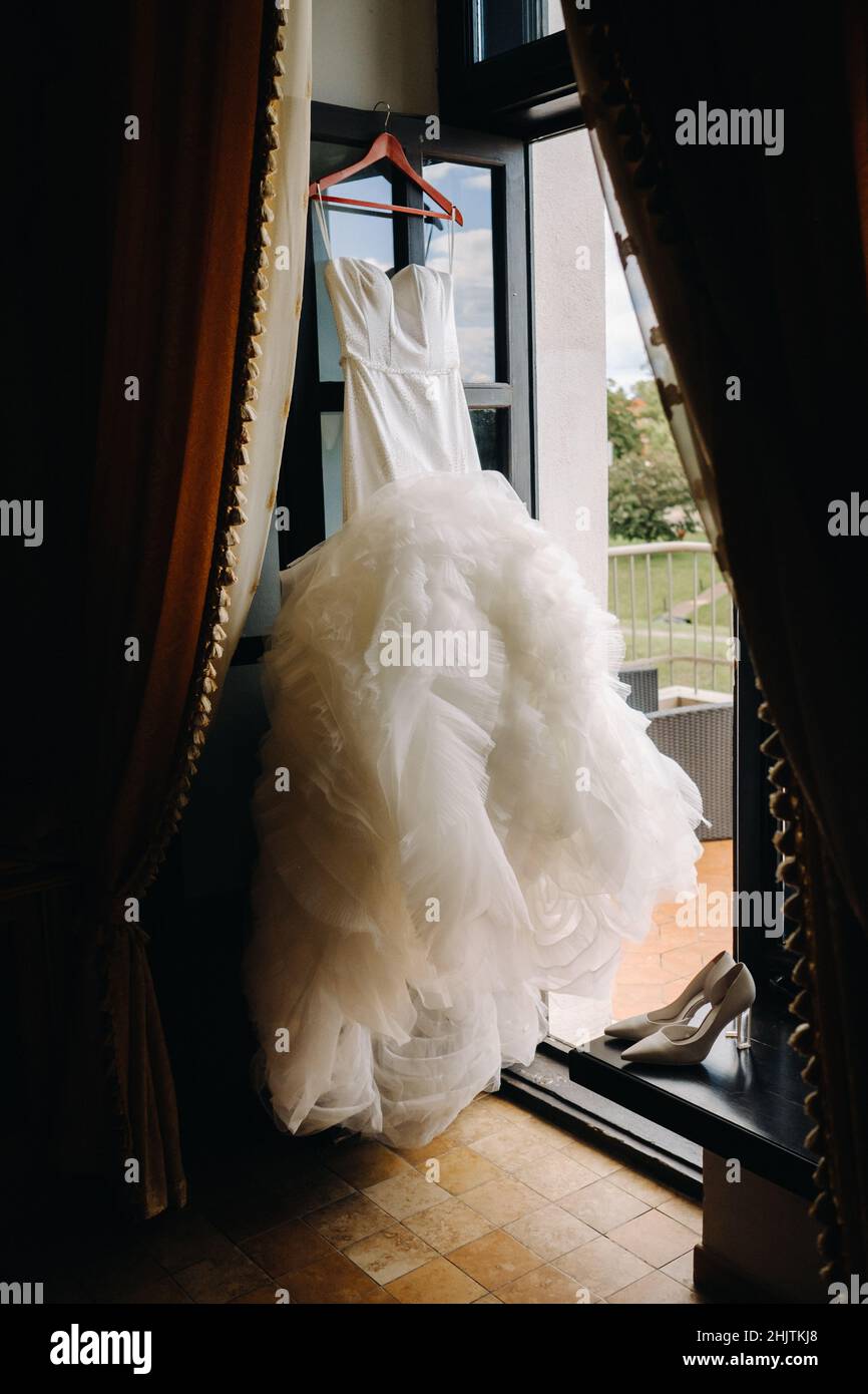 a white wedding dress hangs on a hanger near the window Stock Photo - Alamy