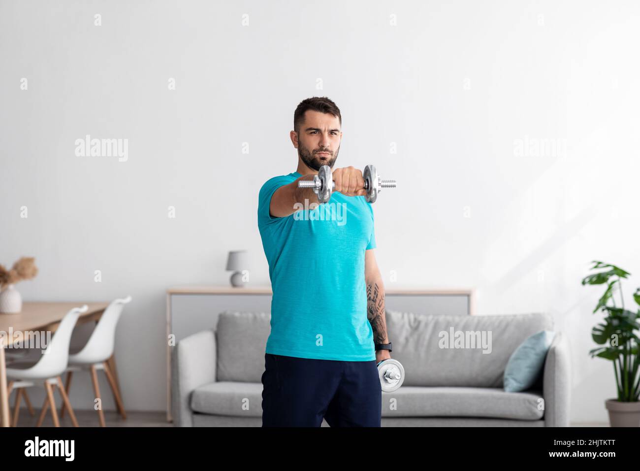 Serious millennial european man athlete in blue t-shirt raises dumbbell in minimalist living room interior Stock Photo