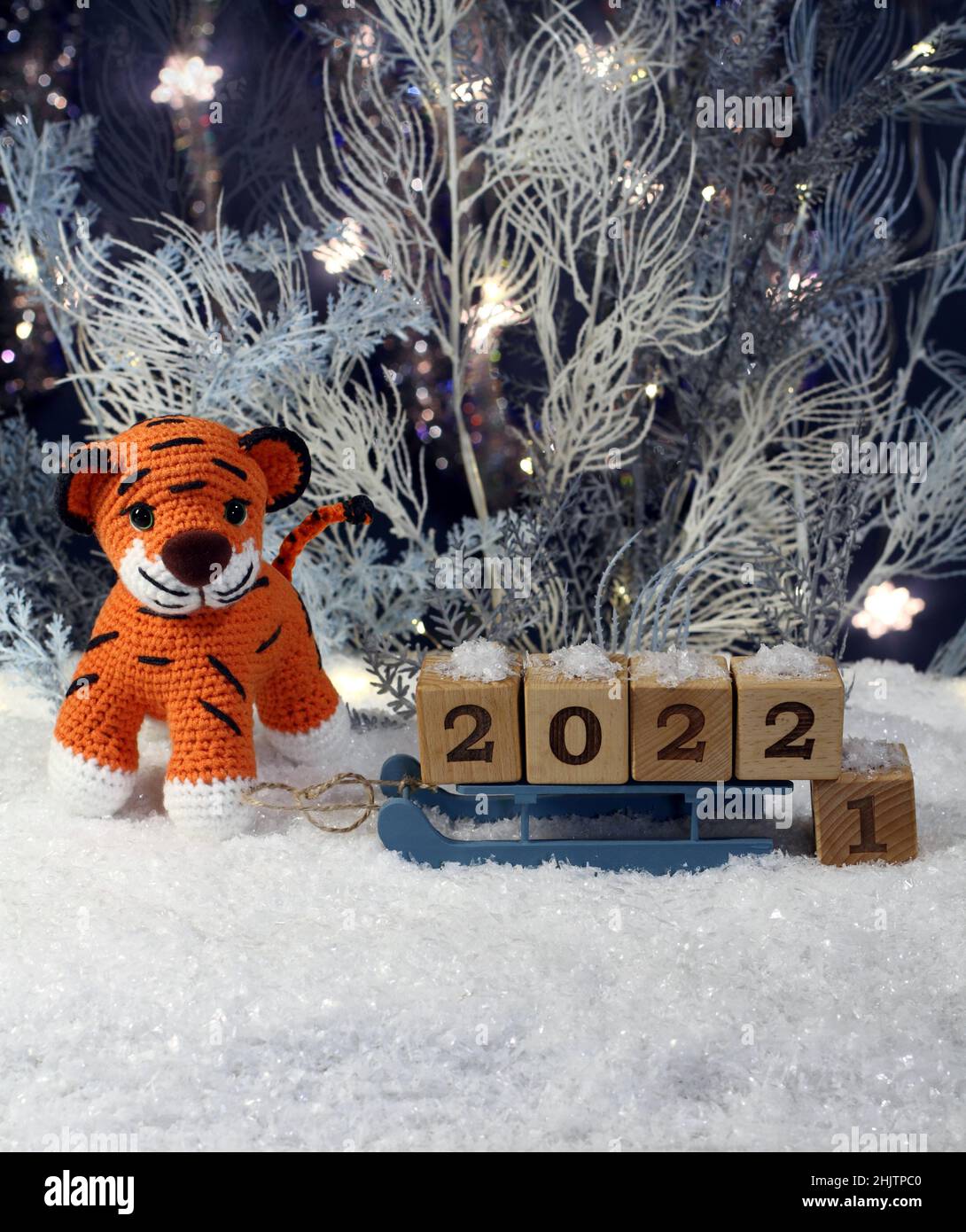 A knitted toy tiger stands on artificial snow and a toy sledge with wooden cubes with the numbers 2022 stands next to him against the backdrop of blue Stock Photo