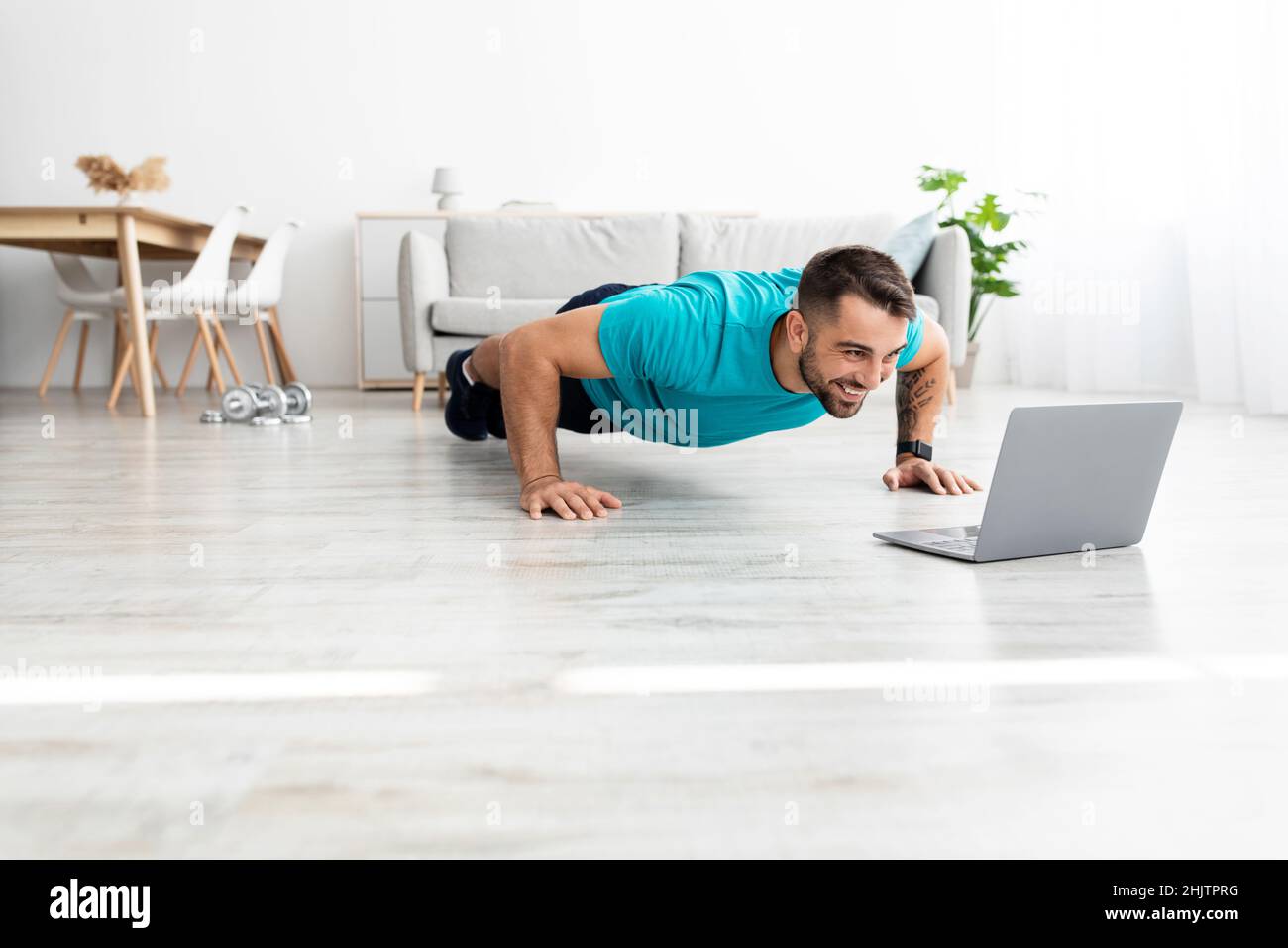 Smiling millennial european male doing push ups and watching online lesson on pc in minimalist living room Stock Photo