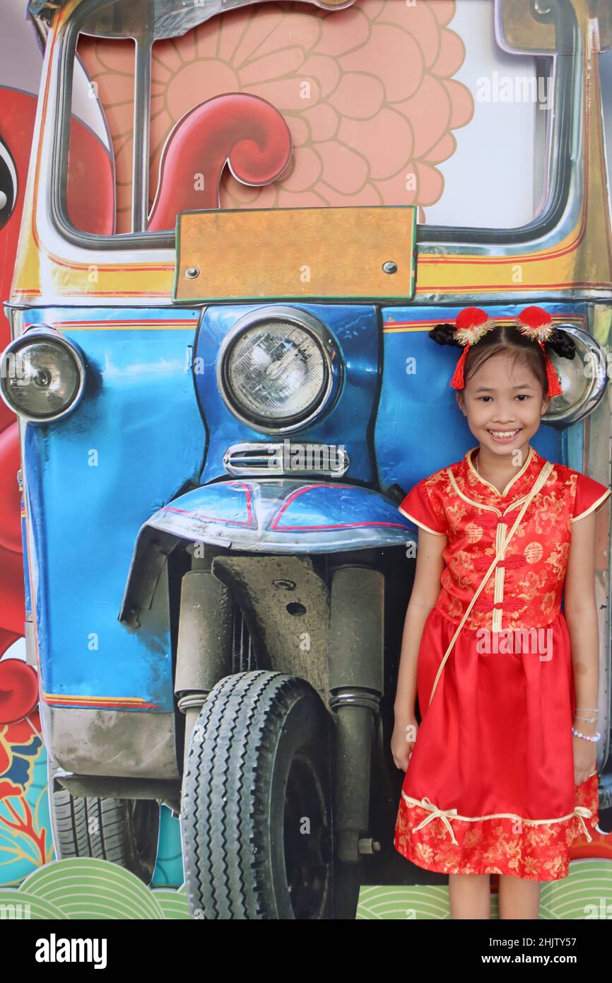 Young Thai Girl wears a Chinese dress and smiles for the camera in front of a mural of a tuk-tuk, Chinatown, Bangkok, Thailand Stock Photo