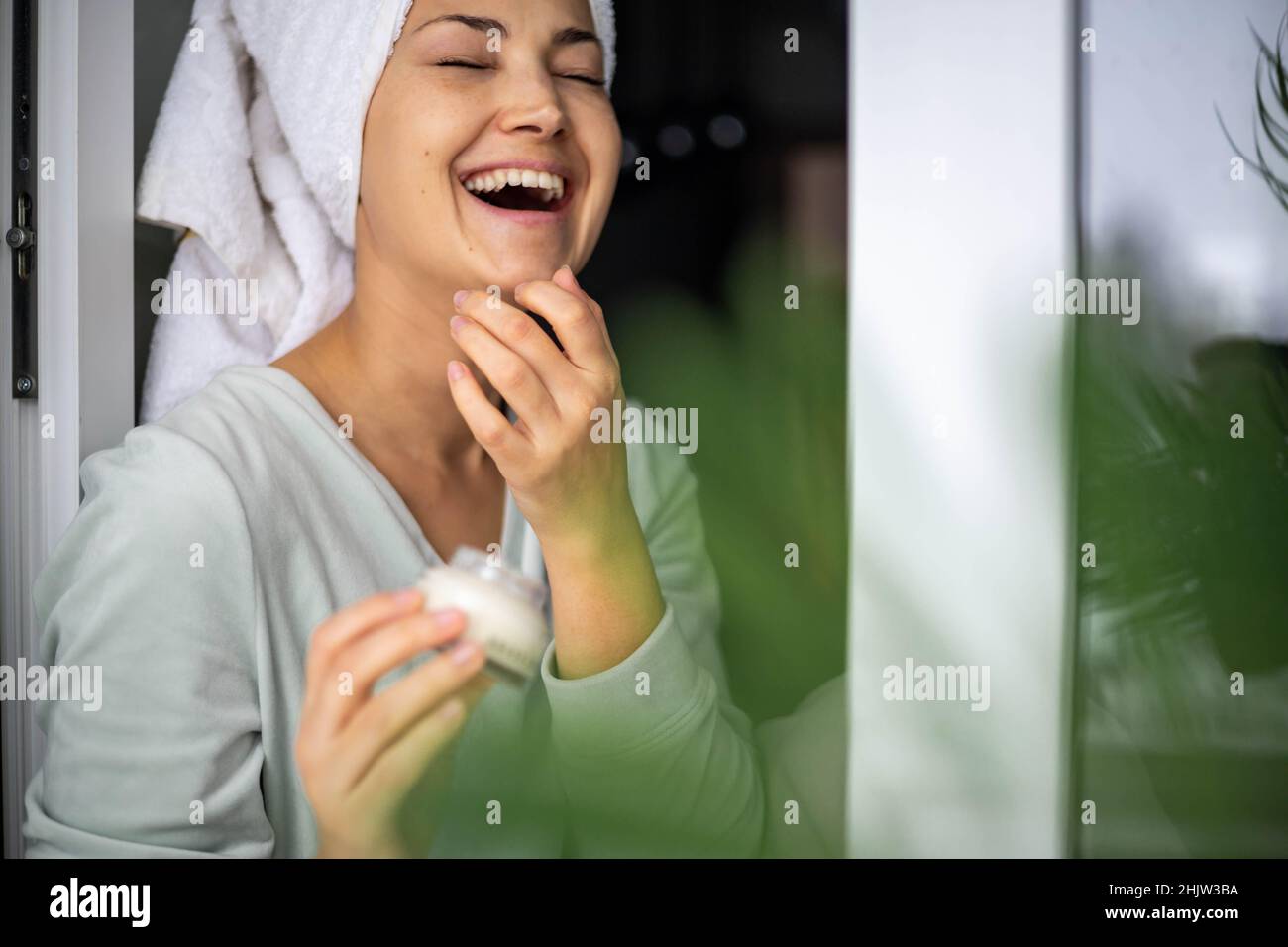 Happy domestic woman applying anti aging cream from jar on face looking at mirror Stock Photo