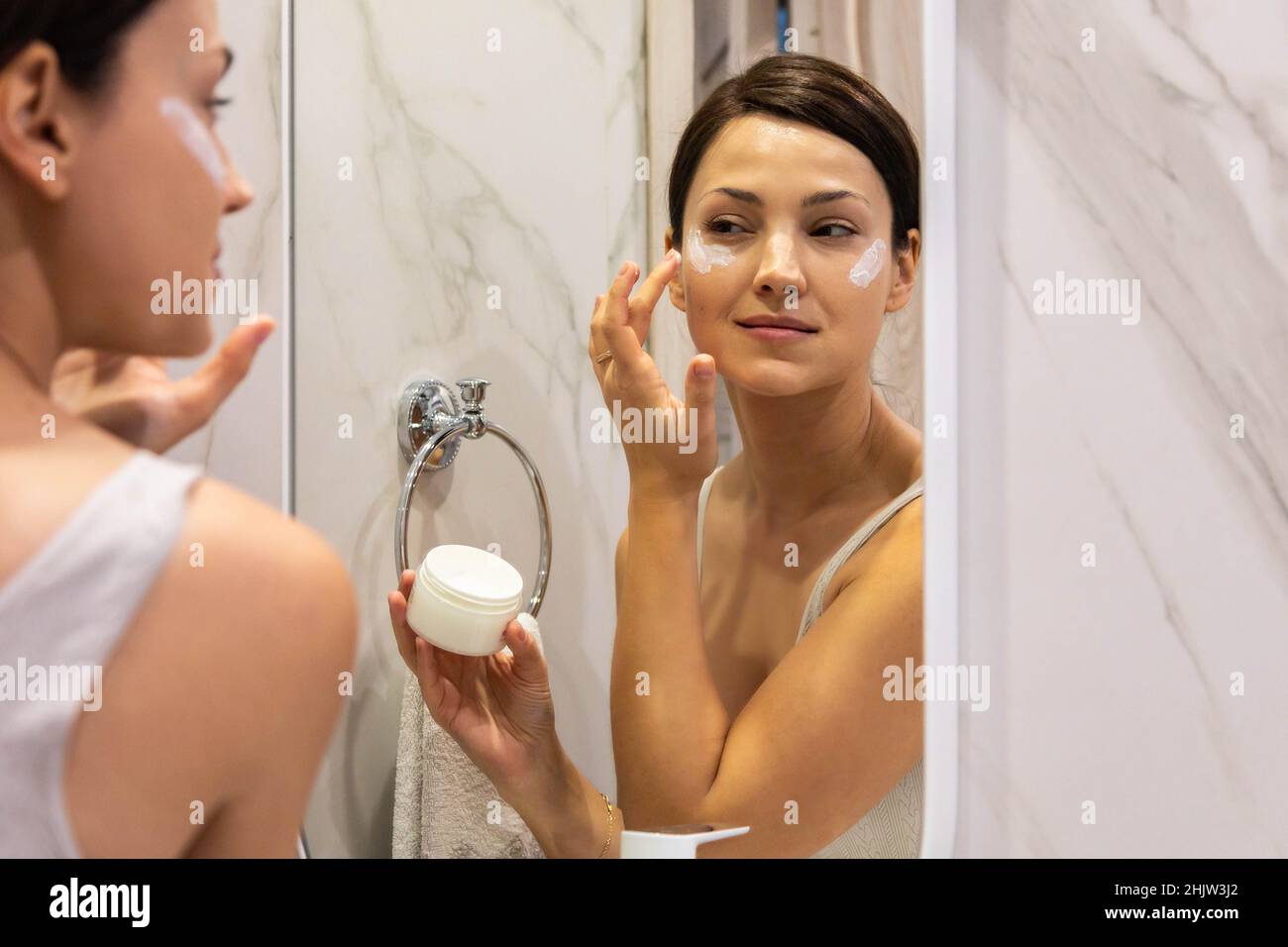 Happy domestic woman applying anti aging cream from jar on face looking at mirror in bathroom Stock Photo