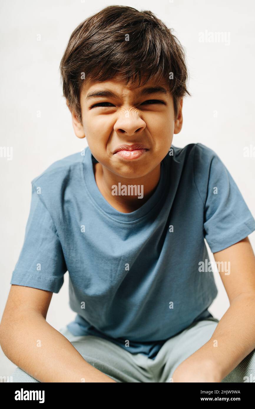 Grimacing indian boy sitting with wrinkled tense face over white  background. He wears simple cotton shirt and pants. Indoors Stock Photo -  Alamy