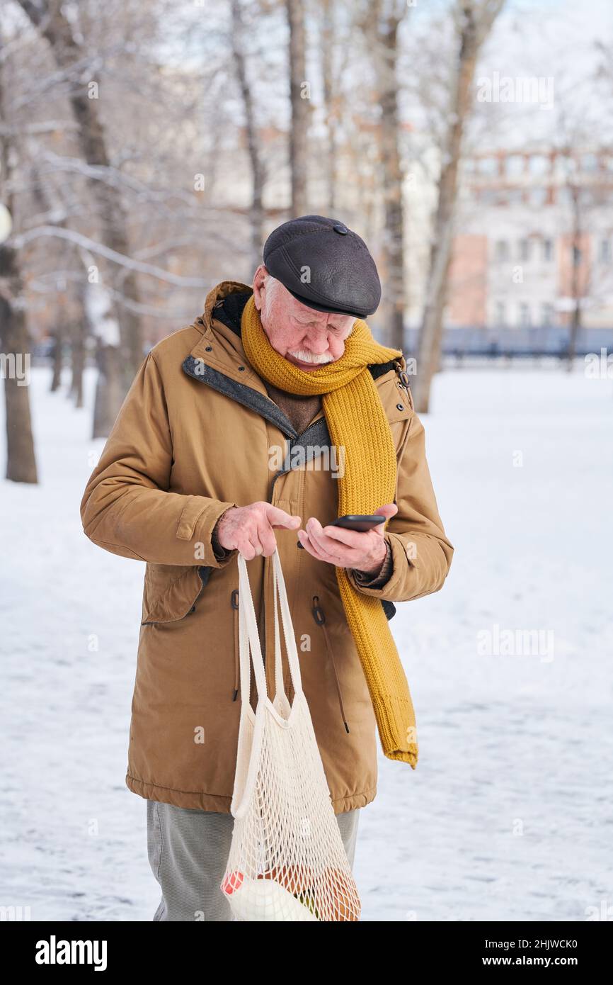 Elderly man in warm clothing holding bag with grocery and using his mobile phone while walking along the street after shopping Stock Photo