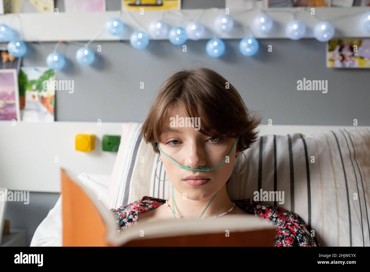 Close-up of sick teenage girl with oxygen tube in her nose reading a book while lying on bed at hospital ward Stock Photo