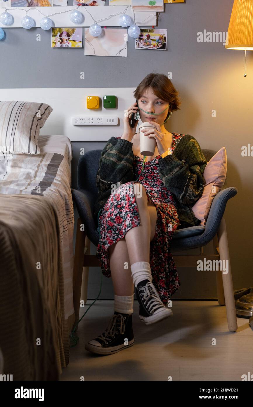 Sick teenage girl sitting on armchair and drinking milk cocktail while talking on mobile phone at hospital ward Stock Photo