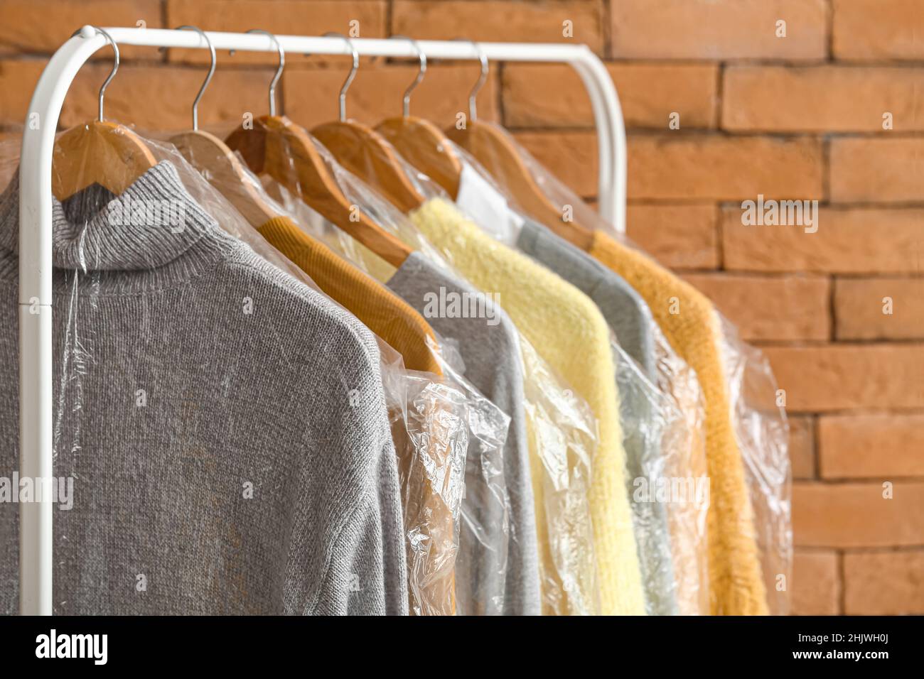 Rack with clean sweaters in plastic bags near brick wall Stock Photo