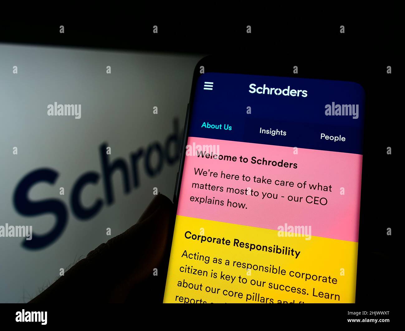 Person holding cellphone with webpage of British asset management company Schroders plc on screen with logo. Focus on center of phone display. Stock Photo