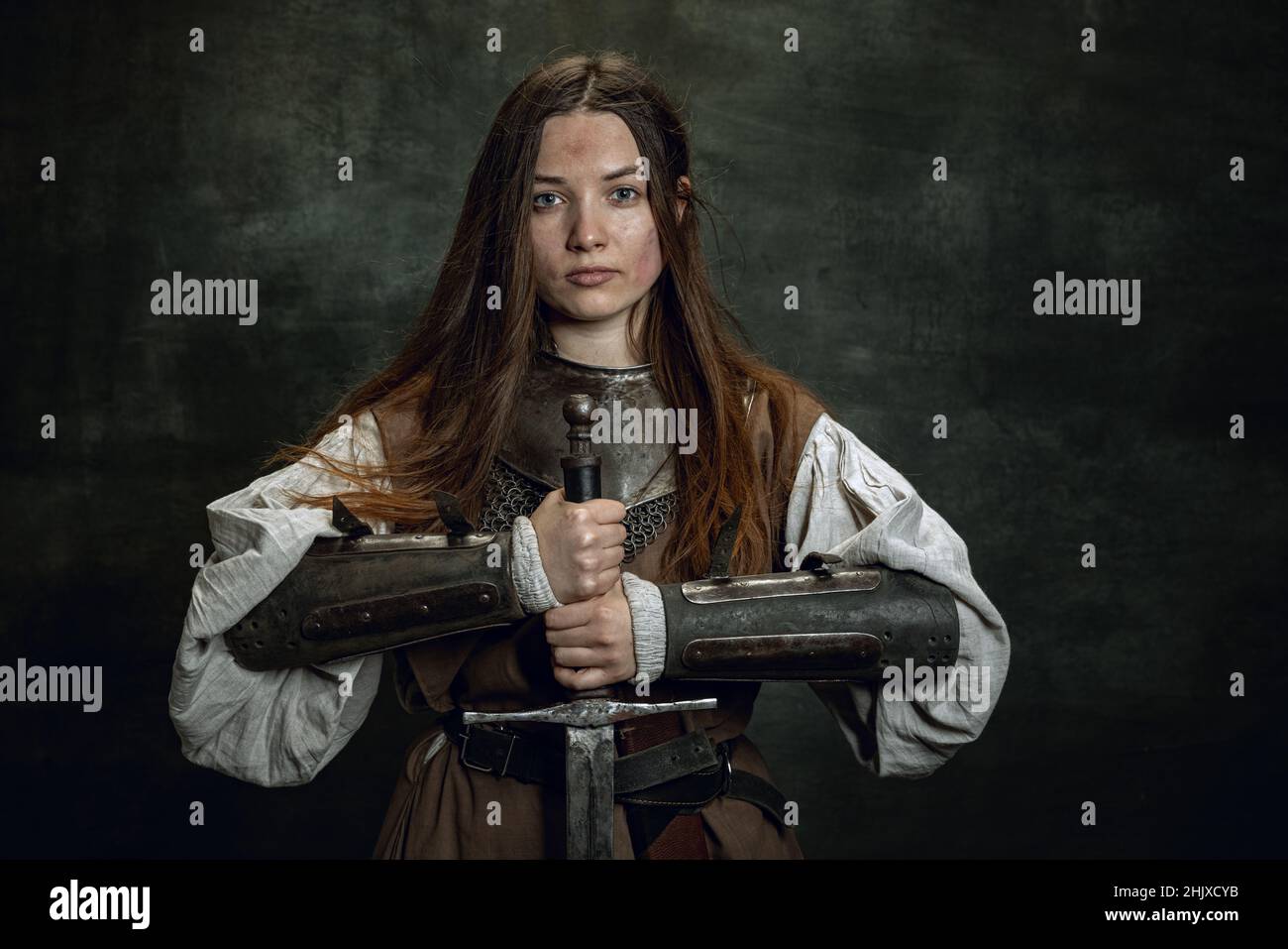 Vintage portrait of adorable woman, medieval female warrior or knight with dirty wounded face looking at camera isolated over dark retro background. Stock Photo