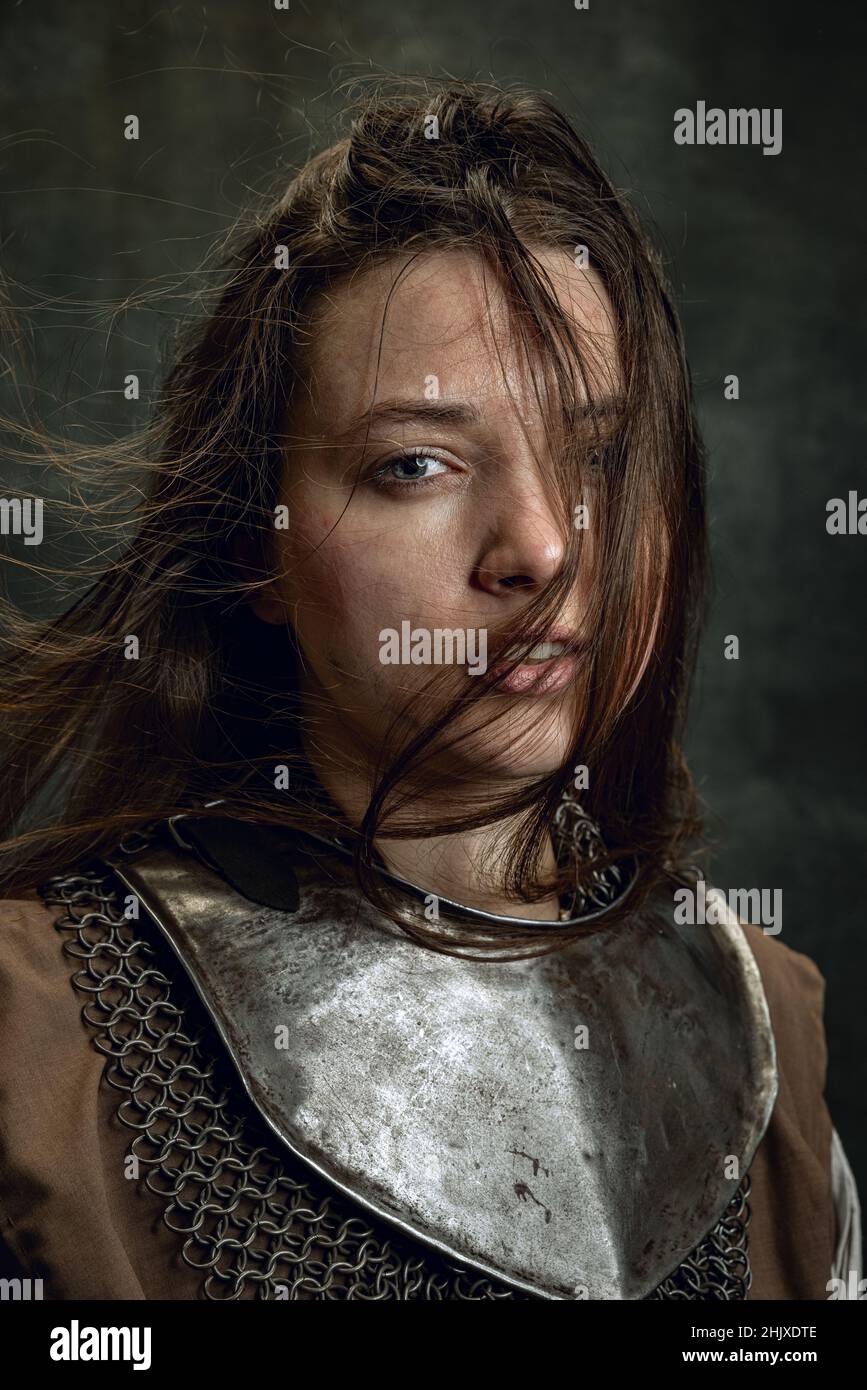 Vintage portrait of adorable woman, medieval female warrior or knight with dirty wounded face looking at camera isolated over dark retro background. Stock Photo