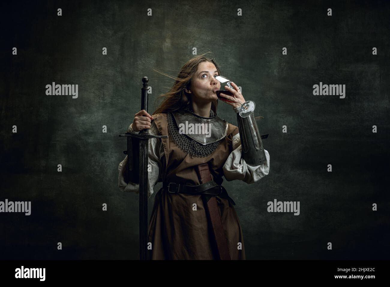 Vintage portrait of adorable woman, medieval female warrior or knight with dirty wounded face tasting wine isolated over dark retro background. Stock Photo
