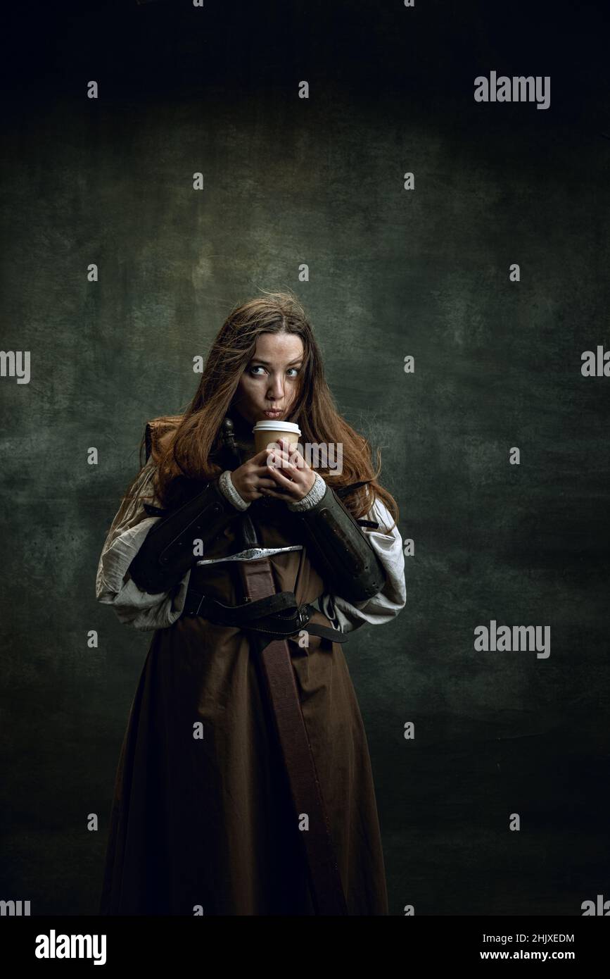 Comparison of eras. Vintage portrait of adorable medieval woman, female warrior or knight with dirty wounded face tasting coffee isolated over dark Stock Photo