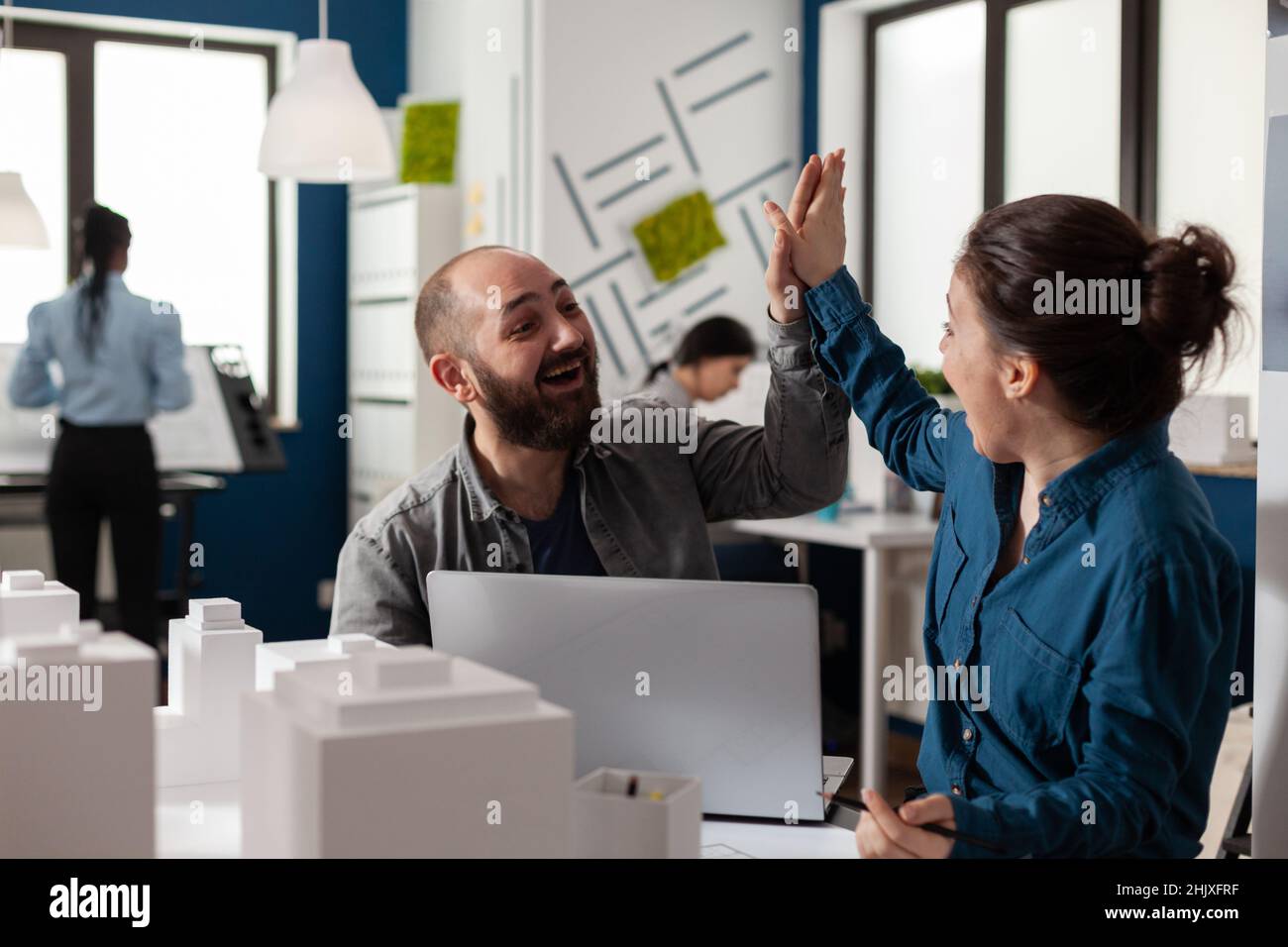 Two architects high fiving sitting at desk in architectural modern office with laptop and foam scale model of residential buildings. Successful happy smiling colleagues celebrating team project. Stock Photo