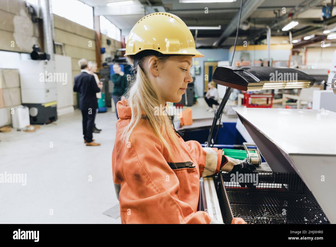 Side view of female blue-collar worker in hardhat using machinery in factory Stock Photo