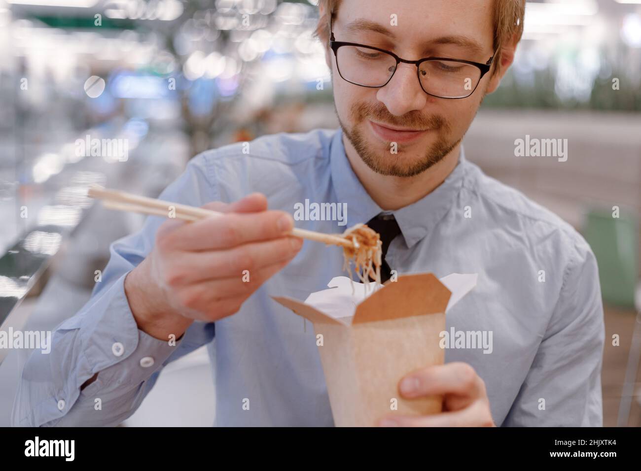 Young businessman with chopsticks eating chinese wok from box on food court. Lunch time. Stock Photo