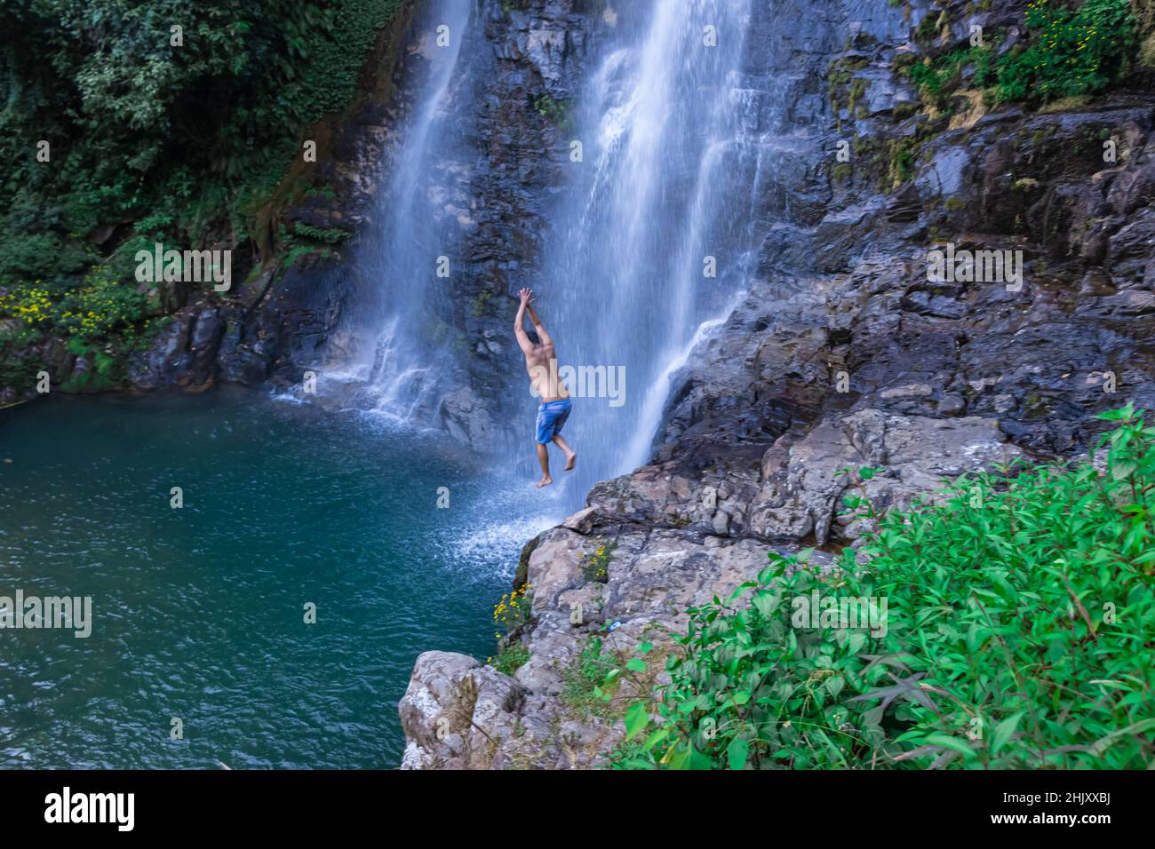young man jumping from cliff at natural waterfall blue water at morning from top angle image is taken at thangsning fall shillong meghalaya india. Stock Photo