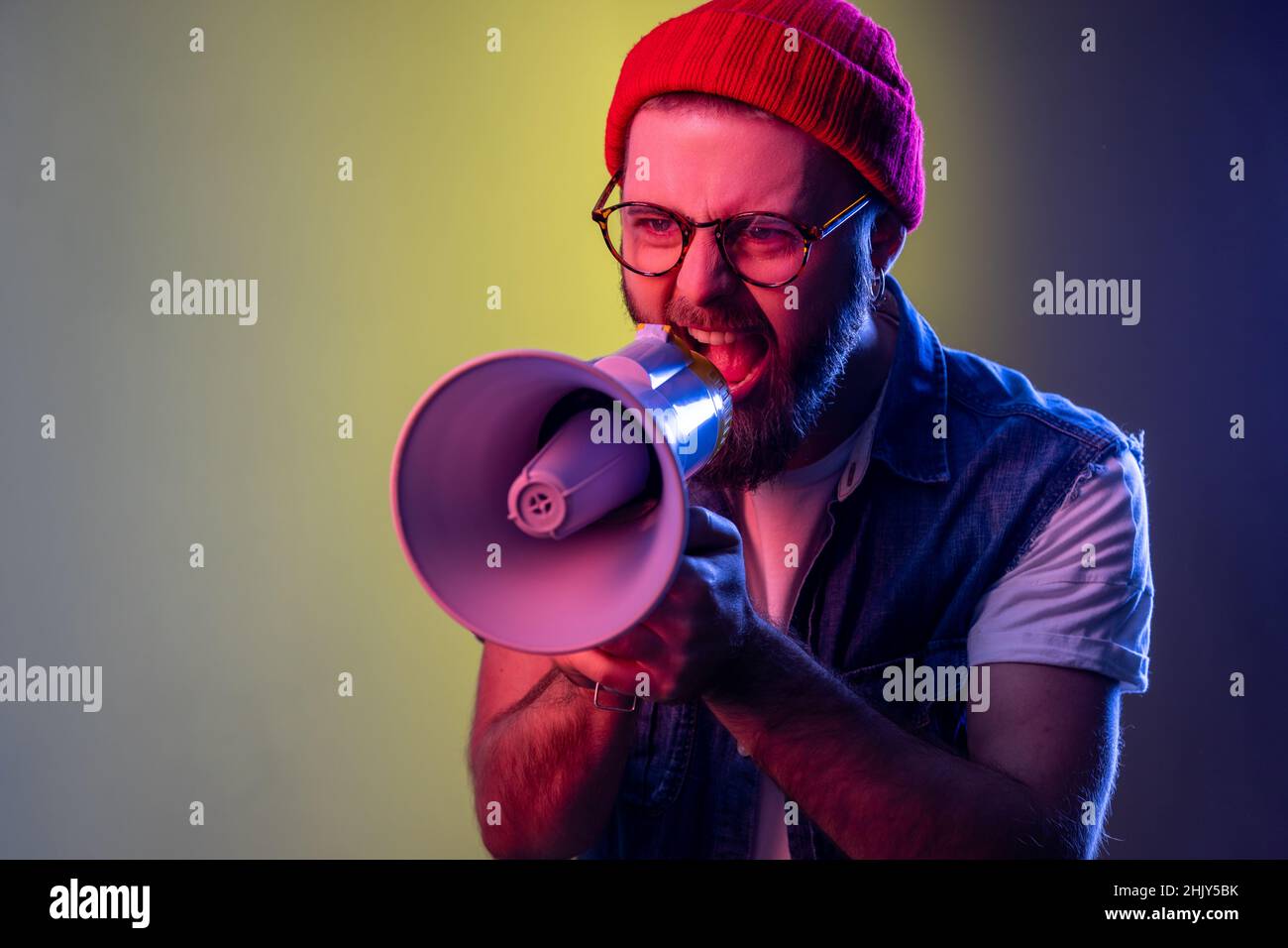 Excited hipster guy screaming loud using megaphone, making announcement, presentation, protesting, wearing beanie hat and denim vest. Indoor studio shot isolated on colorful neon light background. Stock Photo