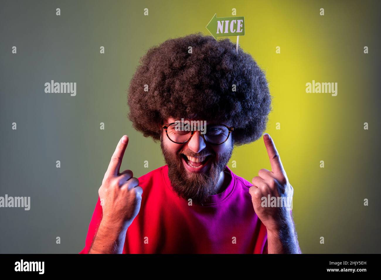 Portrait of positive funny hipster man with Afro hairstyle pointing at party props with nice inscription in his hair, wearing sweatshirt. Indoor studio shot isolated on colorful neon light background. Stock Photo