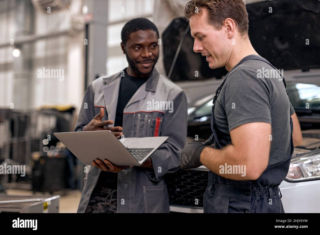 Handsome diverse mechanics in uniform examining car, using laptop while working in auto service. black and caucasian Auto mechanics checking car,servi Stock Photo