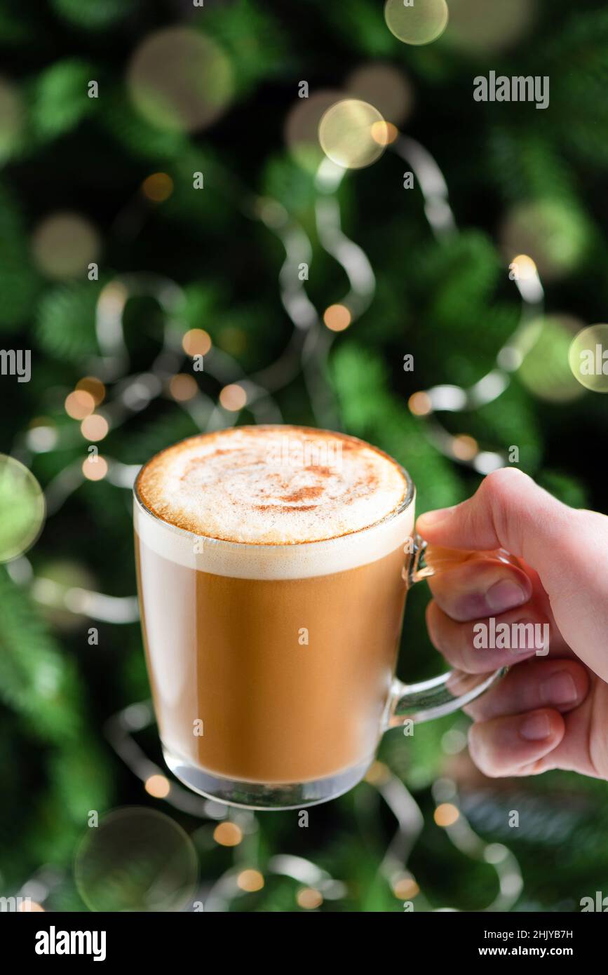 Cup of hot coffee Cappuccino with gingerbread syrup in male hand in front of Christmas tree. Holiday drink Stock Photo