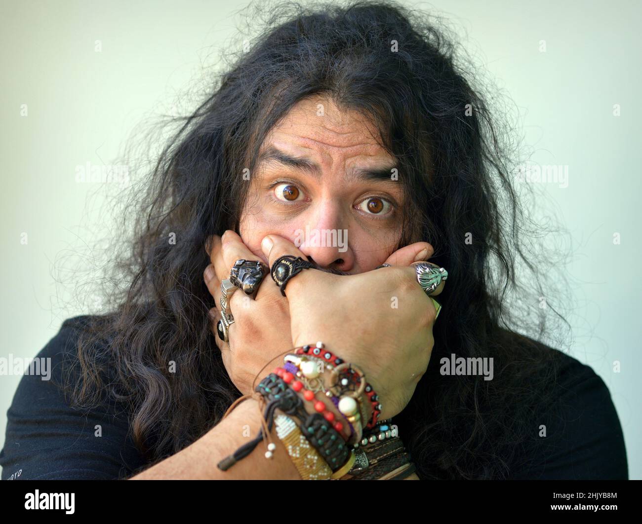 Surprised funny brunette Caucasian young man with big brown eyes and long curly hair covers his mouth with his beringed hands and looks at viewer. Stock Photo