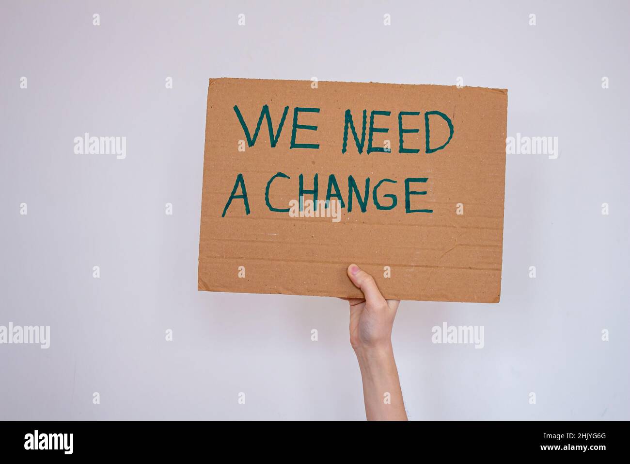 Hand holding cardboard with 'we need a change' inscription over gray wall background. She may be protesting global warming or environmental pollution. Stock Photo