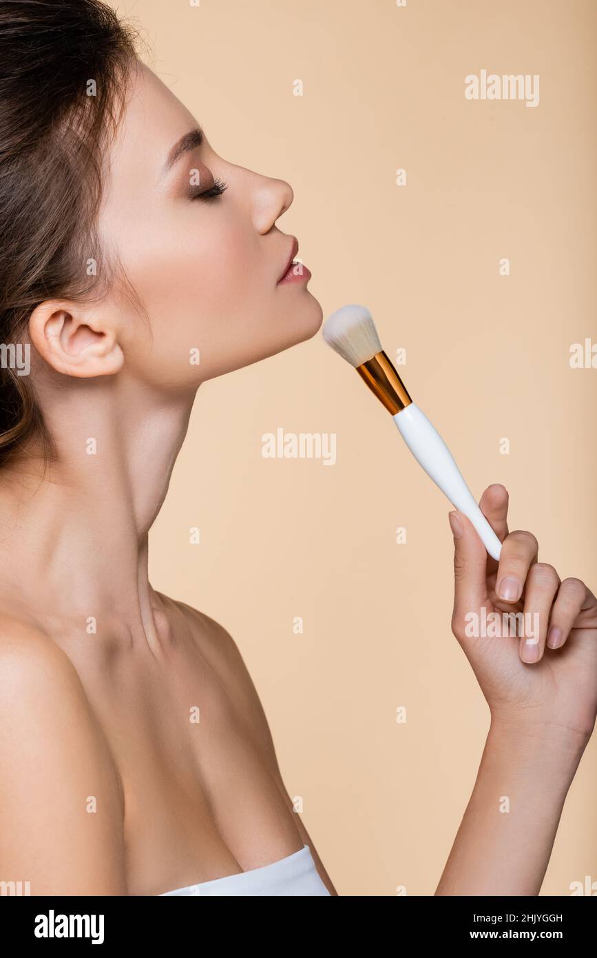 Side view of pretty model with closed eye holding cosmetic brush near chin isolated on beige Stock Photo