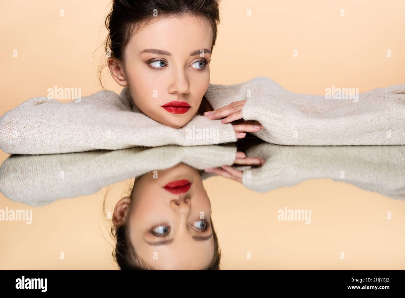 Pretty model with red lips wearing sweater and looking away near mirror isolated on beige Stock Photo