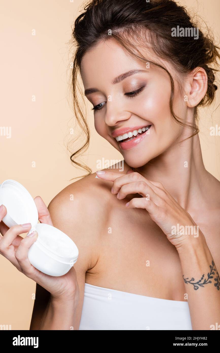 Cheerful woman applying cosmetic cream on shoulder isolated on beige Stock Photo