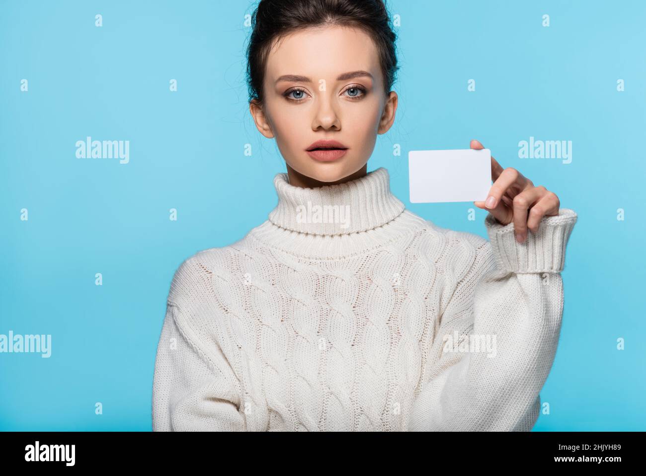 Young woman in knitted sweater holding blank card and looking at camera isolated on blue Stock Photo