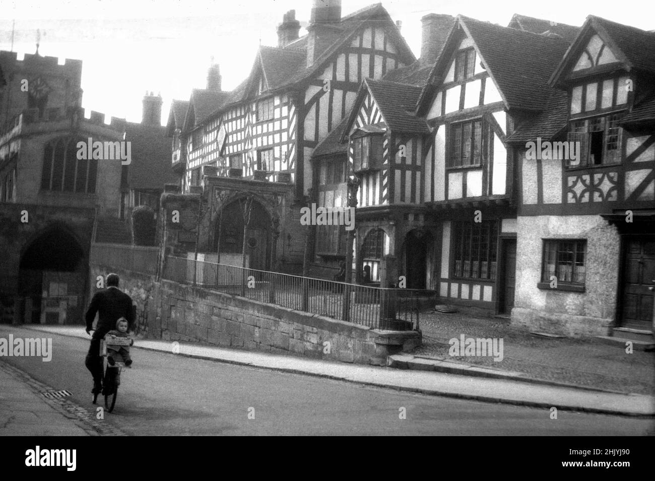1940s, historical view from this time of Lord Leycester's hospital, Warwick, England, UK. Situated next to West Gate on the High Street, Robert Dudley, Earl of Leicester acquired the buildings in 1571, to be used as a hospital for aged or injured soliders under a royal charter from Queen Elizabeth 1. In that era, a hospital did not mean a medical facility, but more of a care or rest home. In the street, we can see a man on a bicycle, with a small child sitting on the back. Stock Photo