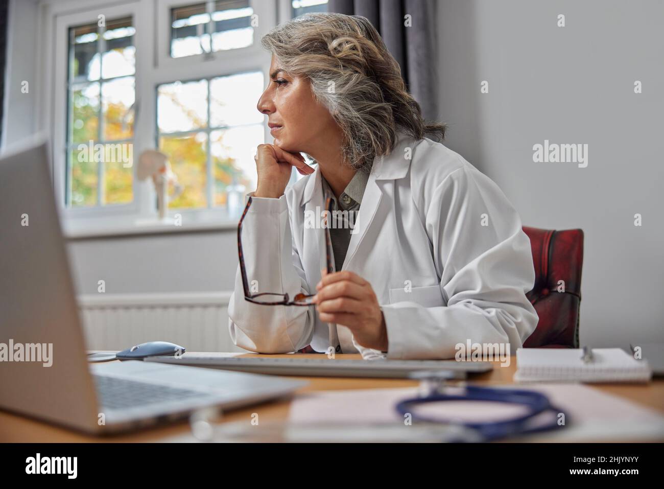 Stressed And Overworked Mature Female Doctor Wearing White Coat At Desk In Doctors Office Stock Photo