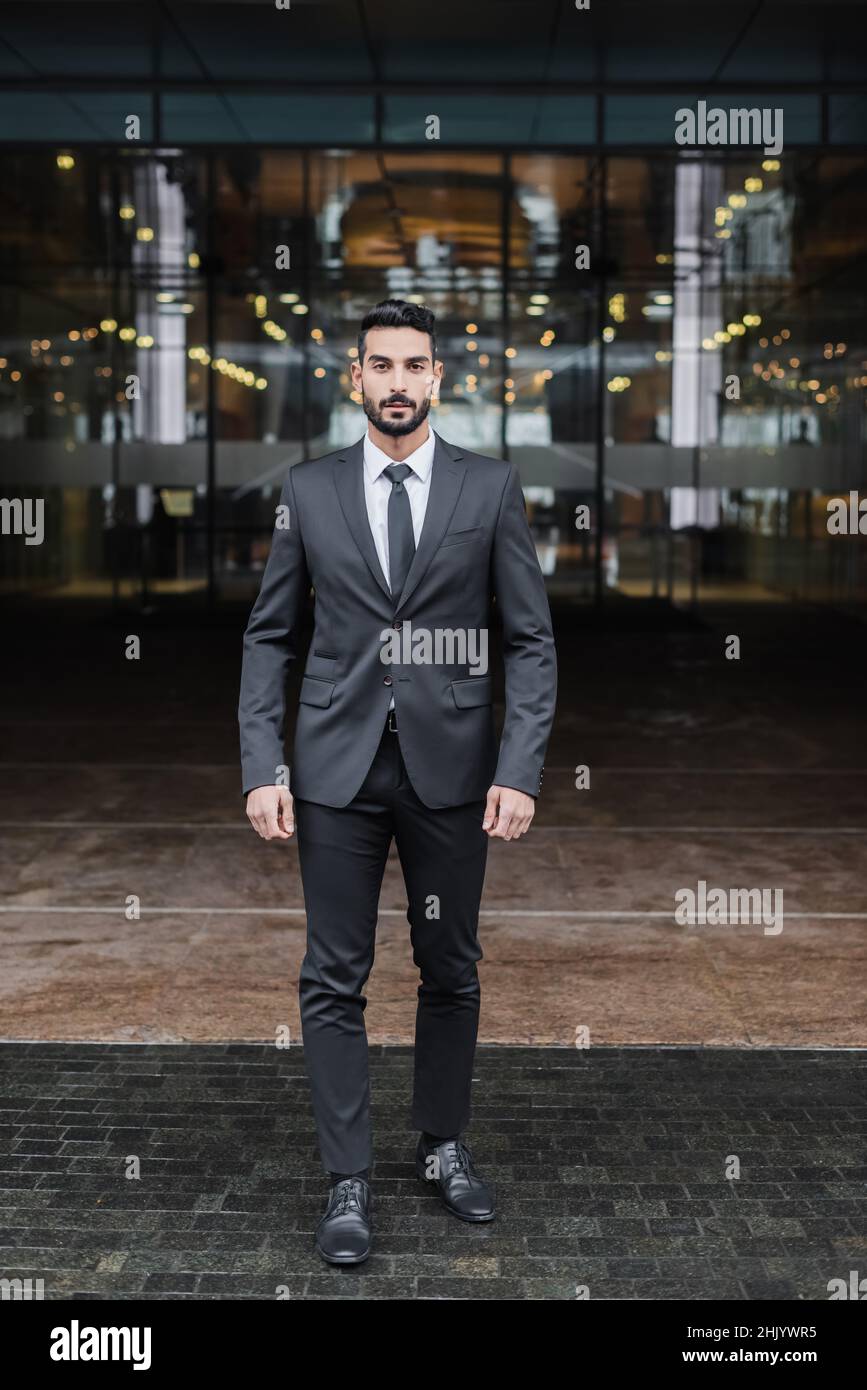 full length view of mixed-race security man looking at camera near blurred hotel building Stock Photo