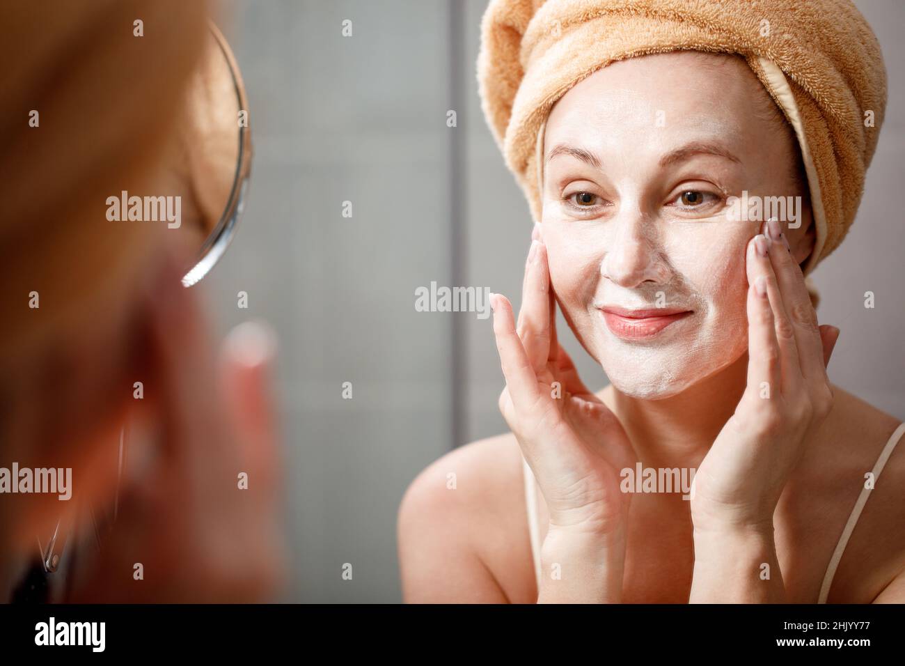 A woman puts a cleansing mask on her face in the bathroom in front of the mirror. Skin care. Cosmetic spa treatments. Stock Photo